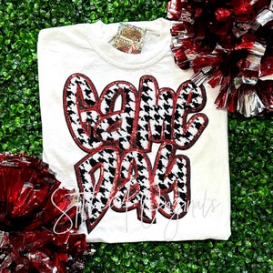 May include: White t-shirt with a black and white houndstooth pattern "Game Day" graphic. The graphic has red accents.