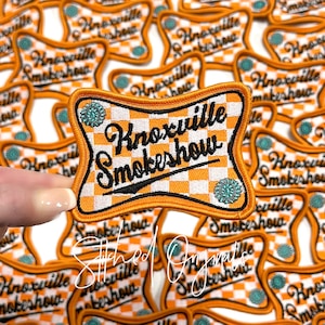 May include: Orange and white checkered iron-on patch with the text "Knoxville Smokeshow" in black cursive lettering. The patch has a white border and two turquoise blue rhinestones.