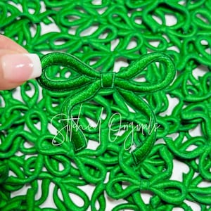 May include: A close-up of a pile of green fabric bows. The bows are all the same size and shape, and they are arranged in a random pattern. The bows are made of a soft, velvety fabric.