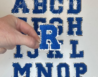 Può includere: Un set di lettere in feltro blu con bordino in glitter argentato. Le lettere sono alte 5,4 cm e sono disposte in ordine alfabetico. La lettera R è tenuta da una mano.