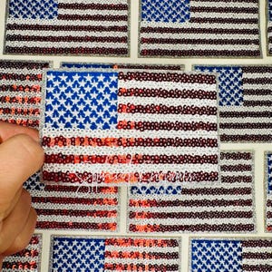 May include: Patches of the American flag, featuring blue star fields and red and white stripes, are displayed. The patches are made of sequins and glitter. The image shows a hand holding one of the patches.