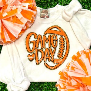 May include: White long-sleeved shirt with an orange and gold glitter design that says "GAME DAY" with a football graphic. The shirt is surrounded by orange and white pom-poms.