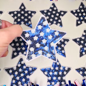 May include: A collection of star-shaped stickers with a patriotic theme. The stickers feature a blue and white sequin design, with a white border. The stars are arranged on a white background.