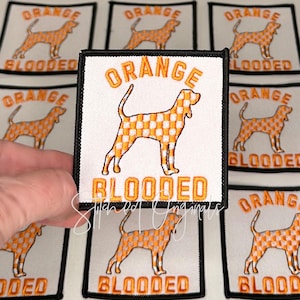 May include: Square white patch with a black border featuring an orange checkered dog silhouette. The words "ORANGE" and "BLOODED" are embroidered in orange above and below the dog. Several similar patches are visible in the background.