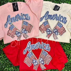 May include: Three sweatshirts with the word "America" in blue sequins and two American flags with a bow in red, white, and blue sequins. The sweatshirts are pink, white, and red.