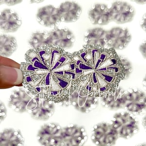 May include: Two silver and purple glitter pom poms with white embroidery. The pom poms are sewn together and are approximately 2 inches in diameter.