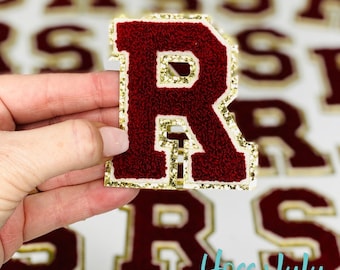 Puede incluir: Un parche con la letra "R" de color granate con ribete dorado brillante. La letra está hecha de un material texturizado y se sostiene frente a un fondo borroso de otros parches de letras. El parche es de Hoss + Lulu.