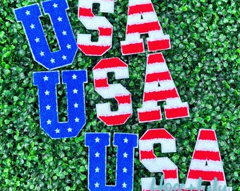 May include: Three sets of felt letter patches spelling out "USA" in red, white, and blue. The letters are in a varsity style font and have a fuzzy texture. The patches are arranged in a stack, with the top set being the smallest and the bottom set being the largest.