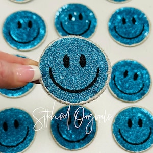 May include: A set of turquoise blue sparkly smiley face iron-on patches. The patches have a silver outline and black stitching. The patches are perfect for adding a fun and quirky touch to clothing, bags, or other accessories.