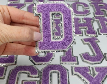 Puede incluir: Parches de letras de fieltro morado con ribete de purpurina plateada. La letra D se sostiene en una mano. Las letras están dispuestas en una cuadrícula.