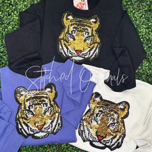 May include: Four sweatshirts in black, periwinkle, and white, each featuring a sequined tiger face design. The tiger's face is gold, black, and white with a red nose. The sweatshirts are displayed on a green background.