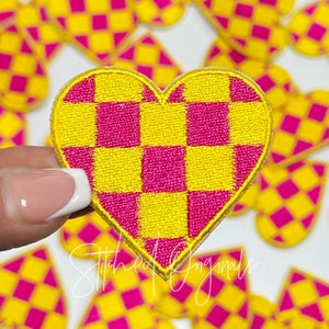 May include: A pink and yellow checkered heart-shaped patch. The patch is made of fabric and has a stitched edge.
