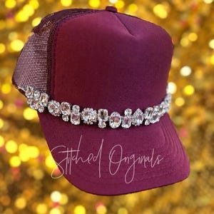 May include: A deep burgundy trucker hat featuring a structured foam front panel and a matching mesh back. A horizontal band of clear, multifaceted rhinestones in various shapes and sizes is attached across the front. The text Stitched Originals is written in white cursive script across the lower portion of the hat.