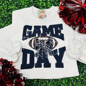 May include: White sweatshirt with the text "GAME DAY" in black glitter letters. A football with a black and silver bow is centered between the words.