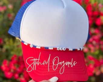May include: Red, white, and blue trucker hat with a beaded trim that spells out "USA". The hat has a white front panel and a red bill. The back of the hat is blue mesh. The hat is held in a hand and the words "Stitched Originals" are written on the bill.
