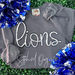 May include: Gray sweatshirt with the word "lions" spelled out in silver sequins. The sweatshirt has a crew neck and long sleeves. The word "Stitched Originals" is printed in white on the bottom of the sweatshirt.