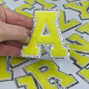 May include: Yellow felt letter patches with a silver glitter trim. The letter A is being held in a hand. The letters are in a variety of sizes.