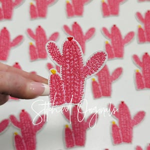 May include: Embroidered pink cactus patch with white border. The cactus has a textured design with a small yellow flower and a red heart. The text "Stitched Originals" is in the image.