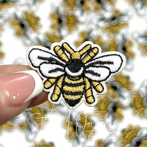 May include: Embroidered bee patch with a black and yellow striped body, white wings, and gold accents. The patch is held in a hand, with a blurred background of similar patches. The text "Stitched Originals" is at the bottom.