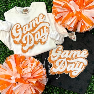 May include: Two shirts with the text "Game Day" in orange and white glitter on a white and black background. The shirts are surrounded by orange and white pom-poms.