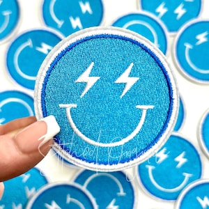 May include: A blue embroidered iron-on patch with a white smiley face and lightning bolts for eyes. The patch is surrounded by a white border.