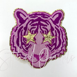 May include: Purple tiger face with gold glitter star eyes. The tiger is outlined in gold glitter.