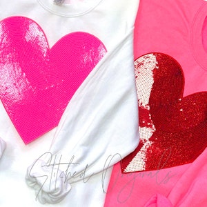 May include: Two sweatshirts with large heart designs. One sweatshirt is white with a pink heart, and the other is pink with a red heart. Both hearts are embellished with sequins.