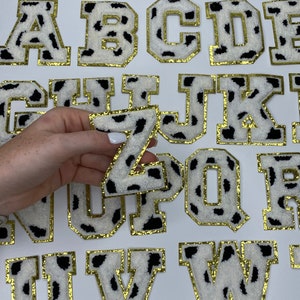 May include: A set of 26 uppercase letters in a cow print pattern with gold trim. The letters are made of a fuzzy material and are perfect for crafting projects.