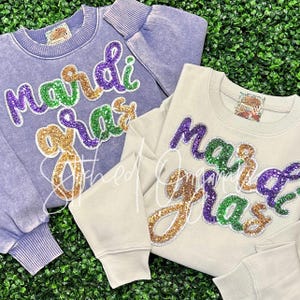 May include: Two sweatshirts with the words "Mardi Gras" in sequined lettering. One is a light purple color, and the other is a light beige color. The lettering is in green, purple, and gold sequins. The sweatshirts have ribbed cuffs and a crew neck.