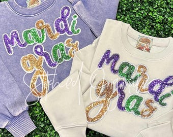 May include: Two sweatshirts with the words "Mardi Gras" in sequined lettering. One is a light purple color, and the other is a light beige color. The lettering is in green, purple, and gold sequins. The sweatshirts have ribbed cuffs and a crew neck.