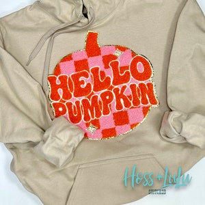 May include: A tan hoodie with a large orange and pink pumpkin patch design that says "Hello Pumpkin" in a retro font. The design is outlined in gold glitter.