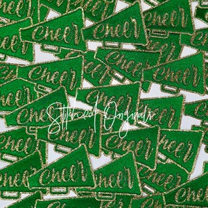 May include: Green megaphone-shaped patches with gold glitter trim and the word "Cheer" in gold glitter script. The patches are scattered across a white surface. The text "Stitched Originals" is also visible.