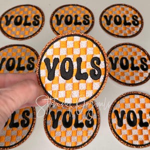 May include: A collection of round embroidered patches with the word "VOLS" in black, set against a checkerboard pattern of orange and white. The patches have a glittery orange border.