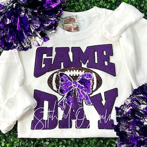 May include: White long-sleeved sweatshirt with purple sequin "GAME DAY" text and a football graphic. The football has a purple sequin bow. The words "Stitched Goals" are below the football. Purple pom-poms are in the background.