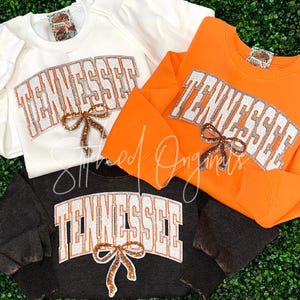 May include: Three sweatshirts with the word "Tennessee" in sparkly silver letters with a brown bow. The sweatshirts are white, orange, and black.