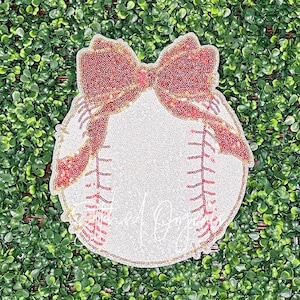 May include: A white baseball with red and gold stitching and a large red bow on top. The baseball is decorated with sequins.