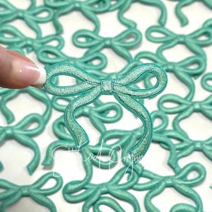 May include: A close-up of a pile of teal blue fabric bows. The bows are all the same size and shape, and they are arranged in a random pattern.