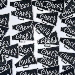 May include: Black megaphone-shaped patches with the word "Cheer" in silver glitter script. The patches are scattered across a white surface, creating a pattern. The patches are likely for cheerleading or spirit wear.