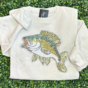 May include: A beige sweatshirt with a large sequined bass fish design. The fish is green, gold, and white.