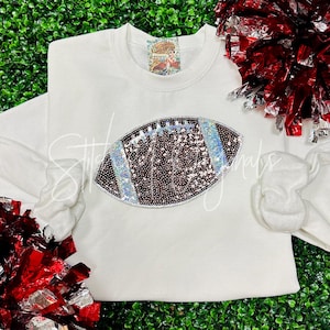 May include: White sweatshirt with a large brown and silver sequined football design.