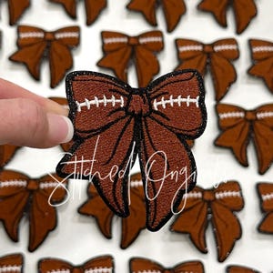 May include: Brown and white football-themed bow with black stitching. The bow is made of fabric and has a glitter finish.