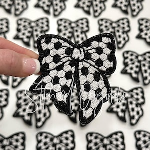 May include: A black and white bow with a soccer ball pattern. The bow is made of fabric and has a glittery finish.