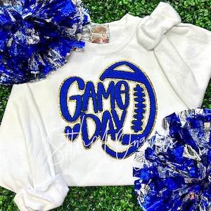 May include: White sweatshirt with a blue and gold "Game Day" football design. The design includes a football shape and the words "Game Day" in blue with gold trim. Blue and silver pom-poms and a white bow are also visible.