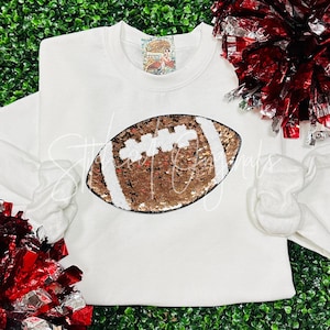 May include: White sweatshirt with a gold and white sequined football design.