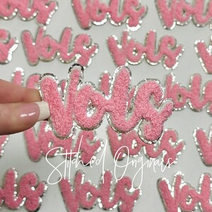 May include: Pink fluffy iron-on patch with the word "Voila" in a cursive font with a silver glitter outline. The patch is surrounded by other similar patches.