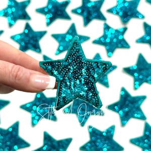 May include: A close-up of a teal blue star-shaped sequin patch. The patch is made of small, shiny sequins that are sewn together to create a star shape. The patch is about 2 inches in diameter.