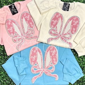 May include: Three long-sleeved shirts with a pink glitter bunny ear design. The shirts are in pink, cream, and light blue.
