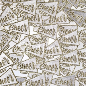 May include: A collection of white and gold glitter "Cheer" megaphone patches. The patches are embroidered with gold glitter lettering and a megaphone design. The patches are scattered across a white background.