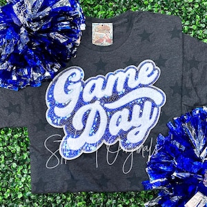 May include: A dark gray t-shirt with a white and blue sequined "Game Day" graphic. The shirt has a star pattern and is surrounded by blue and silver pom-poms.