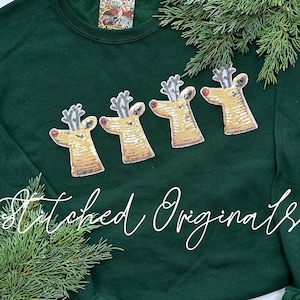 May include: A dark green sweatshirt with four gold sequined reindeer appliques. The reindeer have red noses and black antlers. The sweatshirt has the text "Stitched Originals" in white cursive writing.